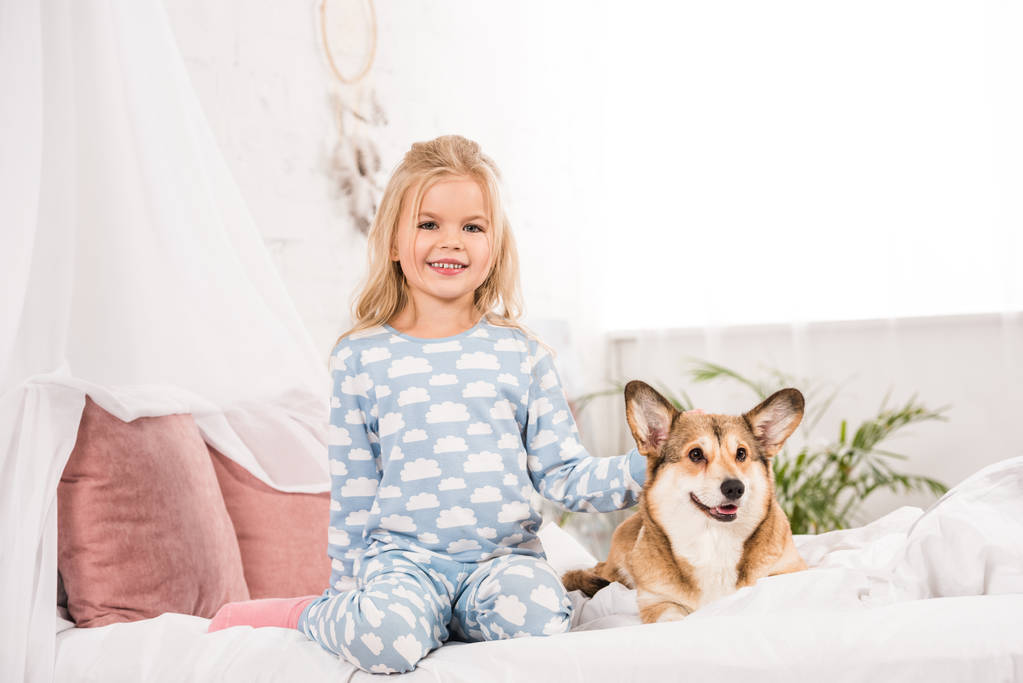 szczęśliwy uśmiechający się dziecko w piżamie siedzi z psem corgi w łóżku - Zdjęcie, obraz