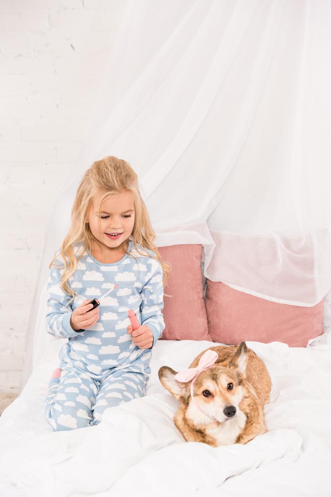 χαριτωμένο παιδί κάθεται στο κρεβάτι με corgi σκυλί και κρατώντας Γκλος Χειλιών  - Φωτογραφία, εικόνα