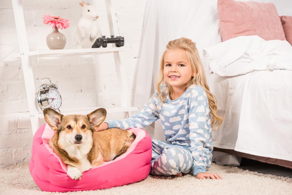 schattig kind in pyjama met welsh corgi dog zitten en kijken naar camera thuis  - Foto, afbeelding