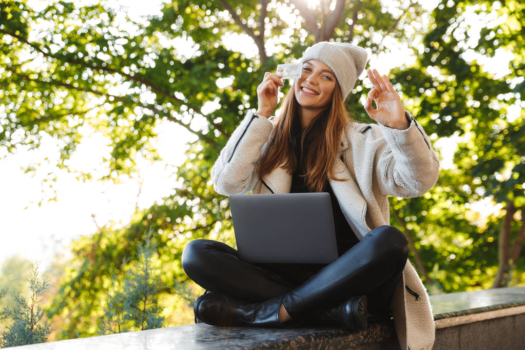 Jovem feliz vestida com casaco de outono e chapéu sentado ao ar livre, usando computador portátil, mostrando cartão de crédito de plástico
 - Foto, Imagem