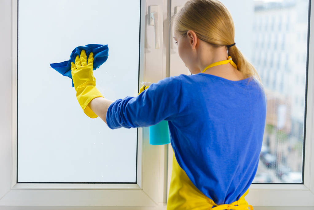 Młoda kobieta w żółtych rękawiczkach czyści szybę w domu szmatą i rozpylanym detergentem. Koncepcja czyszczenia - Zdjęcie, obraz