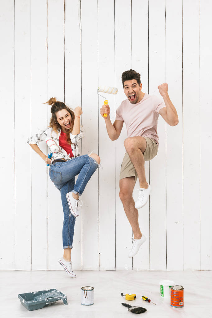Foto de comprimento total do casal alegre homem e mulher pulando enquanto pinta parede branca e faz renovação usando rolos de pintura
 - Foto, Imagem