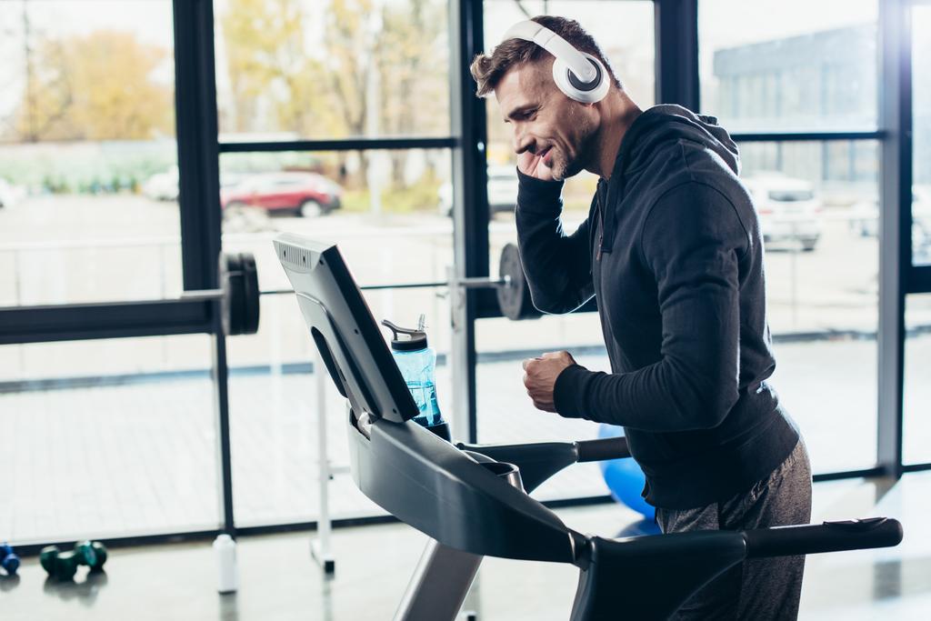 boční pohled pohledný sportovec v hoodie cvičení na běžeckém pásu a poslechu hudby v tělocvičně  - Fotografie, Obrázek