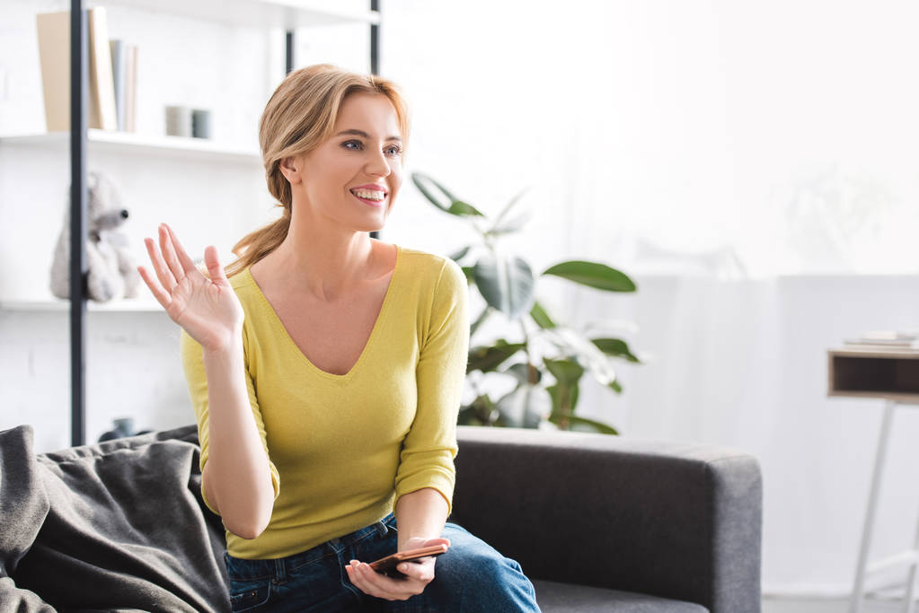 beautiful smiling woman holding smartphone and waving hand while sitting on couch at home - Photo, Image
