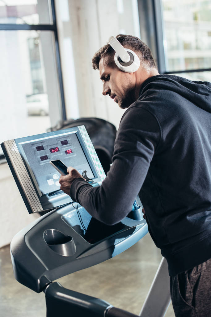 красивый спортсмен в толстовке, тренирующийся на беговой дорожке и пользующийся смартфоном в тренажерном зале
  - Фото, изображение
