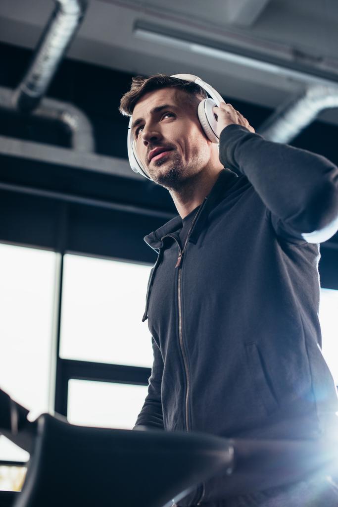 low angle view of handsome sportsman in hoodie exercising on treadmill and listening to music in gym - Photo, Image