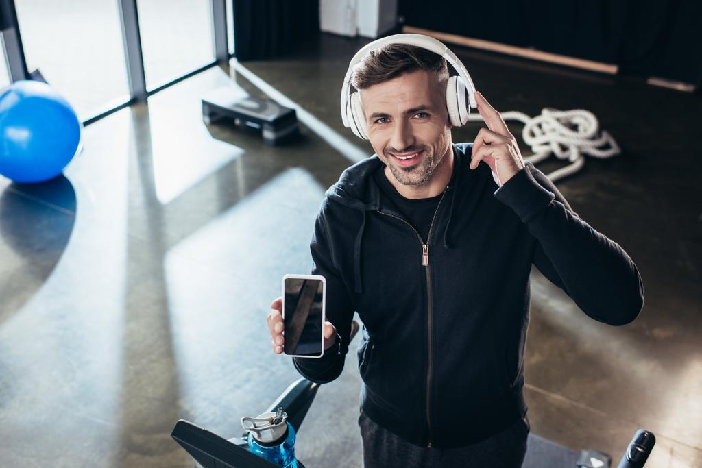 Hochwinkelaufnahme eines lächelnden gutaussehenden Sportlers im Kapuzenpulli, der Smartphone mit leerem Bildschirm auf dem Laufband im Fitnessstudio zeigt  - Foto, Bild