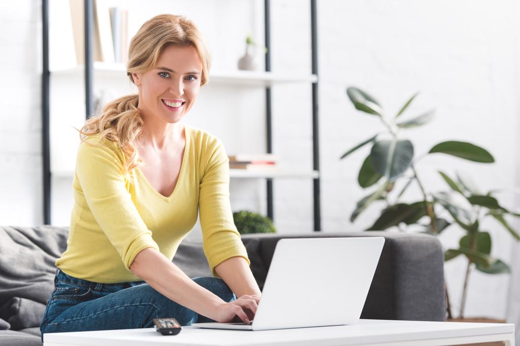aantrekkelijke vrouw met laptop en glimlachend op camera thuis - Foto, afbeelding