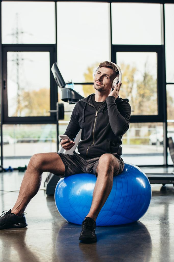 desportista bonito alegre sentado na bola de fitness e ouvir música com smartphone no ginásio
  - Foto, Imagem