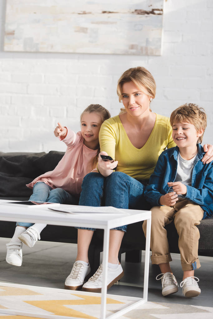 счастливая мама с восхитительно улыбающимися малышами, использующими пульт дистанционного управления и вместе смотрящими телевизор
   - Фото, изображение