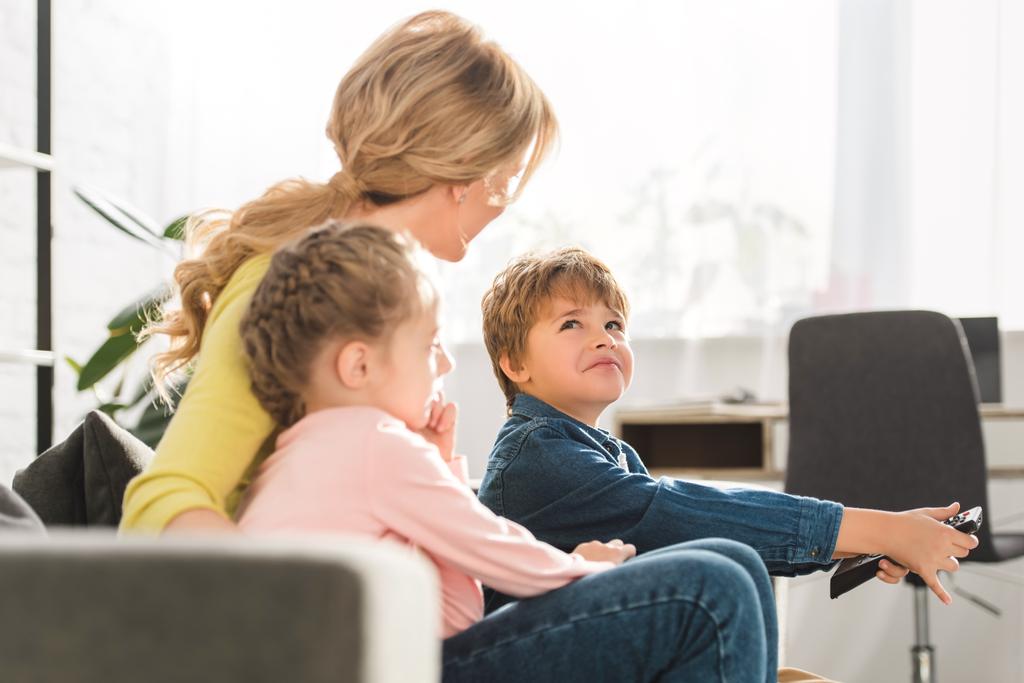 mãe com crianças adoráveis sentado no sofá e usando controlador remoto
 - Foto, Imagem