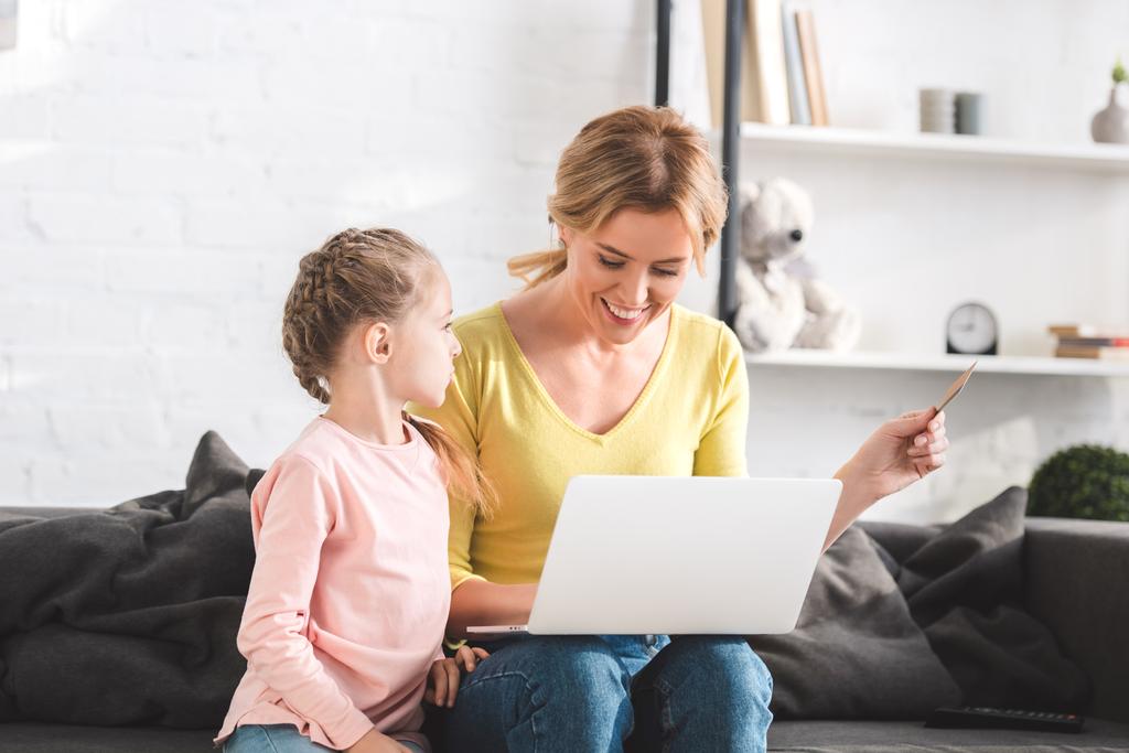 happy mother holding credit card and using laptop with little daughter at home - Photo, Image