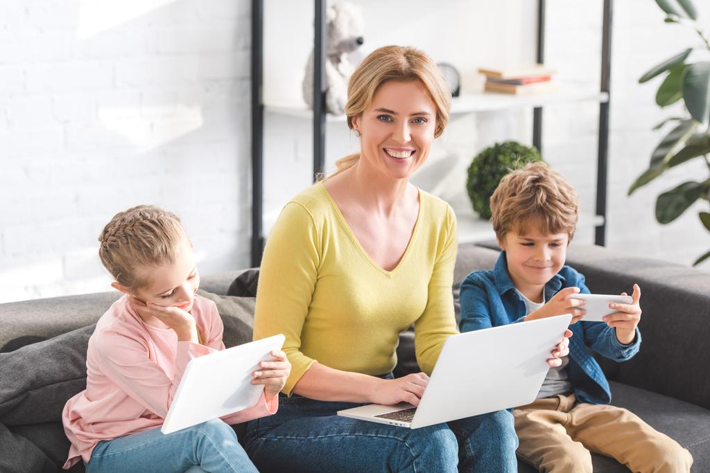 glückliche Mutter mit Laptop und lächelnd in die Kamera, während Kinder digitale Geräte zu Hause benutzen - Foto, Bild