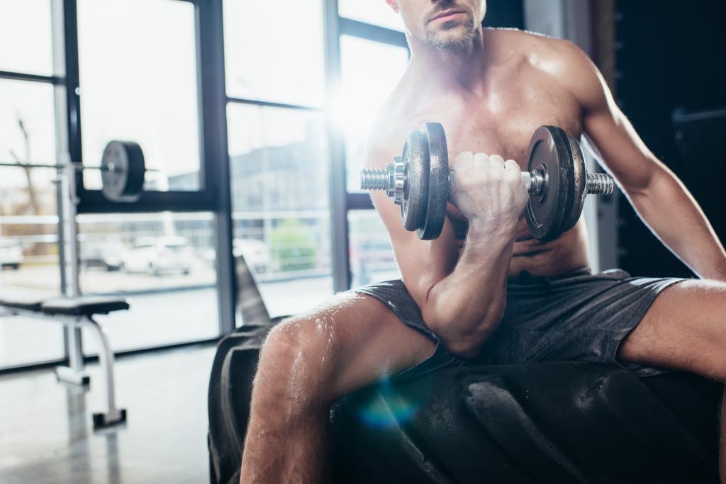 abgeschnittenes Bild eines hemdlosen Sportlers, der auf Reifen sitzt und mit einer Fitnesshantel trainiert - Foto, Bild