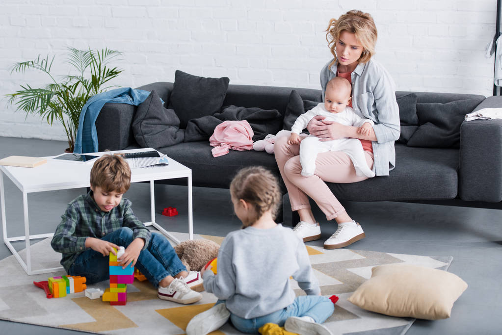 madre cansada con niño pequeño sentado en el sofá y mirando a los hermanos jugando con juguetes
  - Foto, Imagen