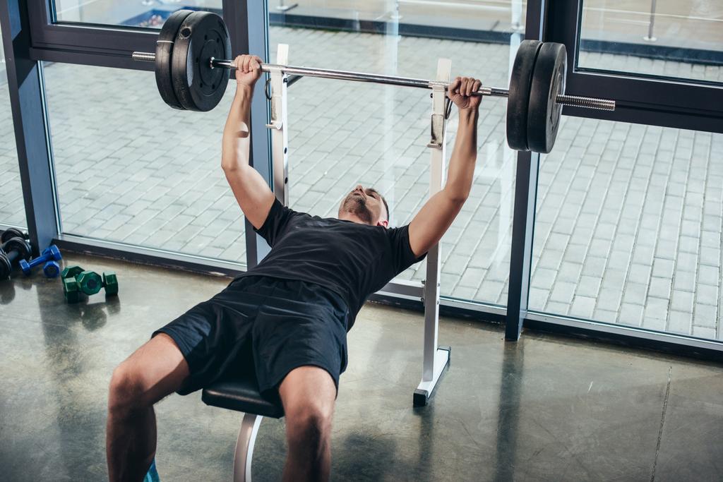 hombre deportivo guapo levantando la barra con pesas mientras está acostado en el banco en el gimnasio
  - Foto, Imagen