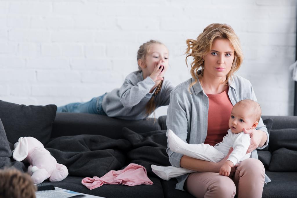 Erschöpfte Mutter hält Säugling und schaut in Kamera, während unartige Tochter hinter ihr spielt - Foto, Bild