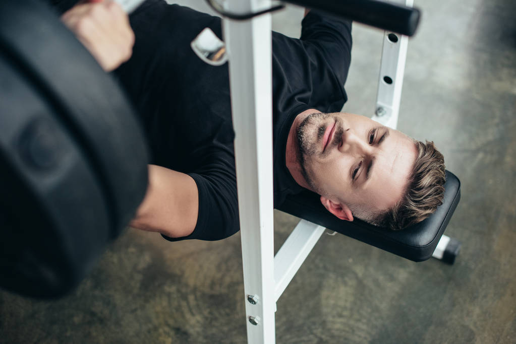 wysoki kąt widzenia przystojny sportowiec podnoszenia sztangi z ciężarkami leżąc na ławce w siłowni  - Zdjęcie, obraz