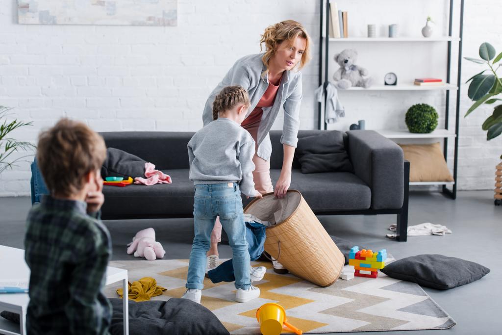 уставшая мать кладет игрушки в корзину, пока непослушные дети играют дома
 - Фото, изображение