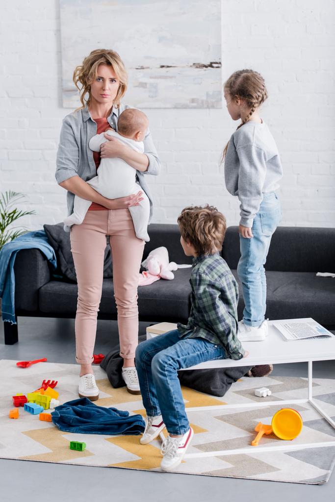 exhausted mother holding infant kid and looking at camera while naughty kids playing in room - Photo, Image