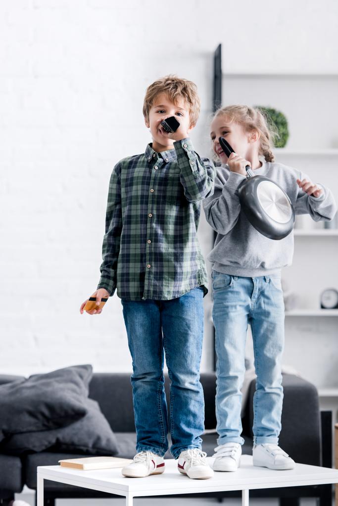 cute brother and sister having fun and singing with remote controller and frying pan - Photo, Image