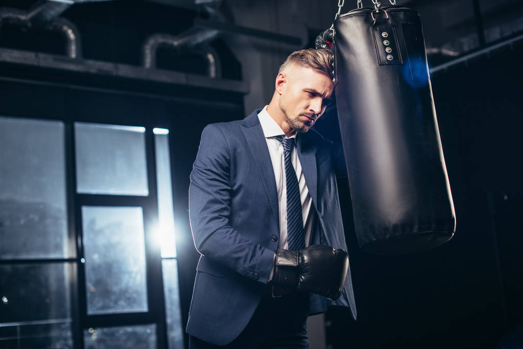 pohledný podnikatel unavený v obleku a Boxerské rukavice, opíraje se o boxovací pytel v tělocvičně - Fotografie, Obrázek
