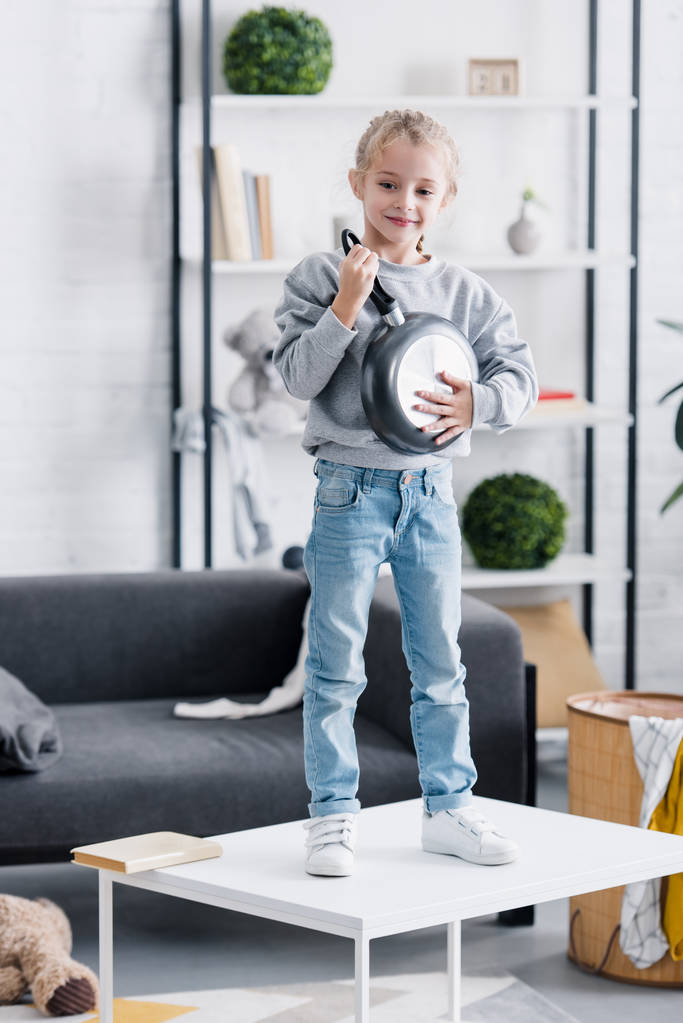 niño feliz de pie en la mesa y jugando con la sartén en casa
 - Foto, imagen