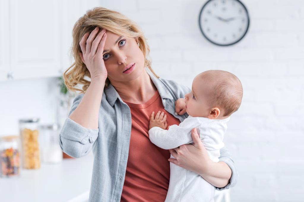 Müde Mutter hält Säugling und steht zu Hause mit der Hand auf der Stirn - Foto, Bild