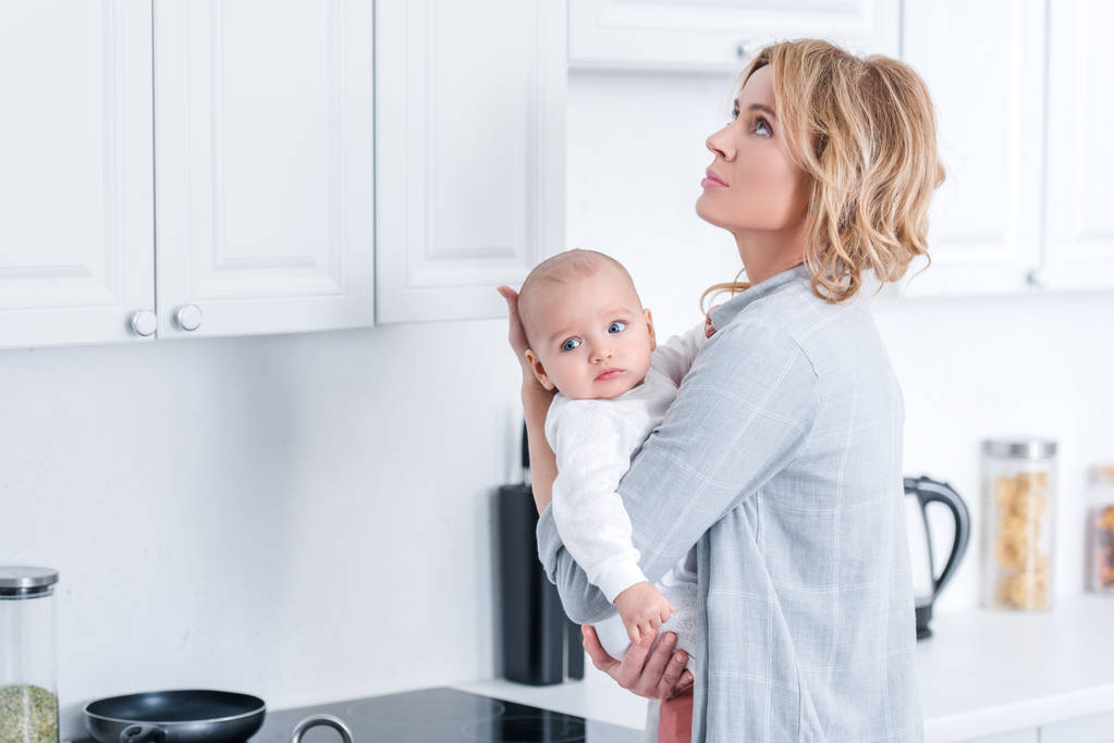 уставшая мать держит младенца ребенка и глядя вверх на кухне
  - Фото, изображение