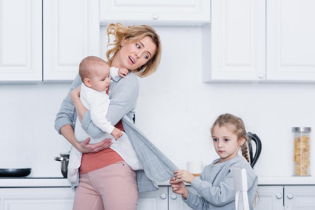 mère épuisée tenant enfant en bas âge tandis que fille coquine tenant son cardigan dans la cuisine
  - Photo, image