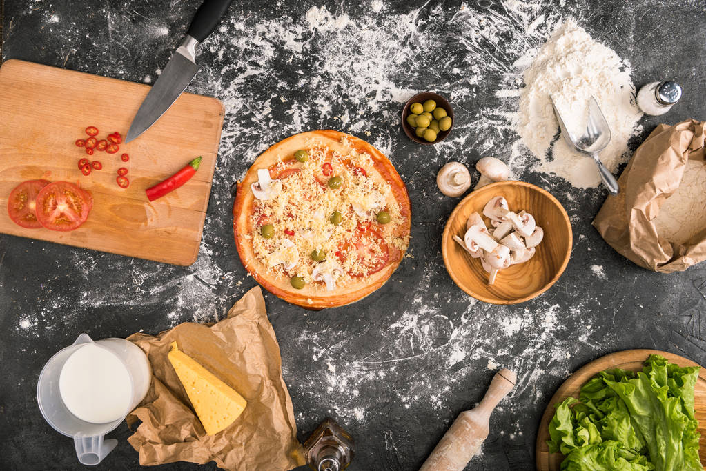 vista superior de pizza cruda, utensilios de cocina e ingredientes sobre fondo gris con harina
  - Foto, imagen