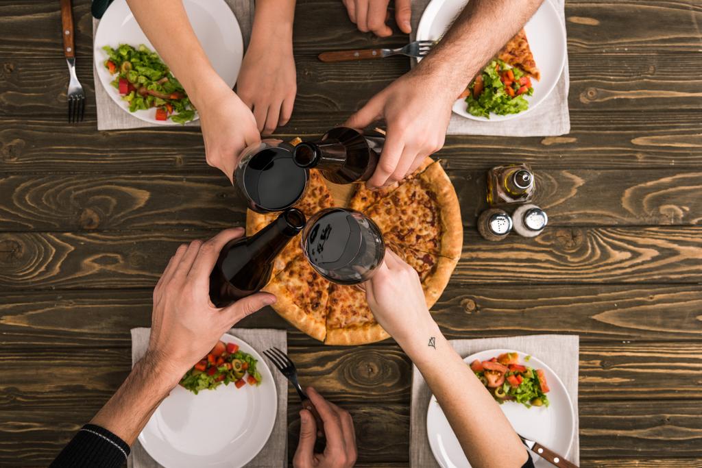 vista cortada de amigos brindar enquanto janta com pizza e saladas na mesa de madeira
 - Foto, Imagem