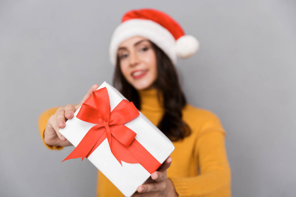 Alegre joven mujer con sombrero de Navidad de pie aislado sobre fondo gris, sosteniendo la caja actual
 - Foto, Imagen