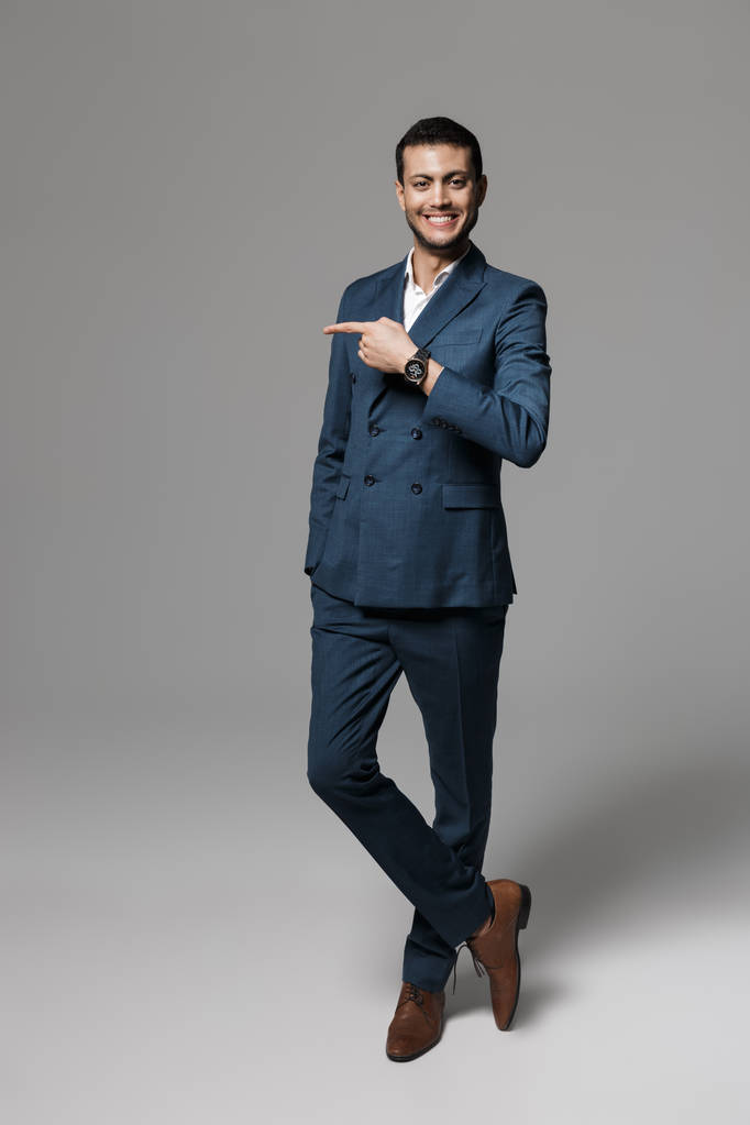 Imagem de comprimento total do atraente empresário árabe 30 anos em terno formal posando na câmera com o dedo de lado isolado sobre fundo cinza
 - Foto, Imagem