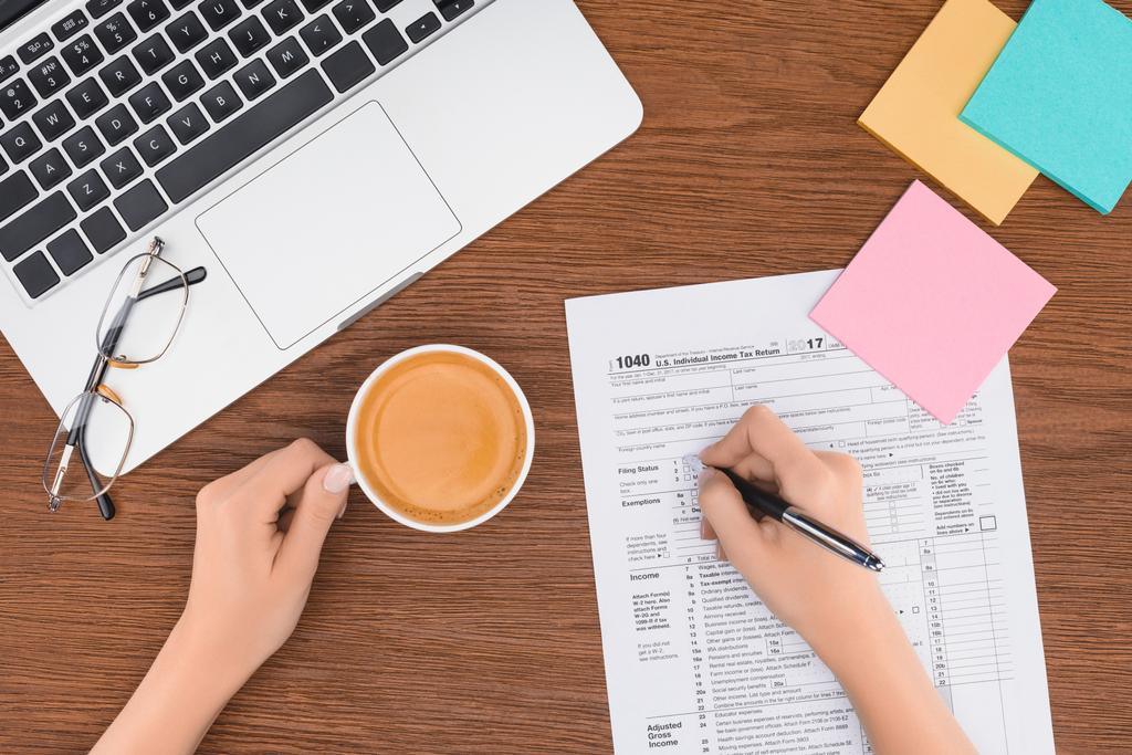 cropped view of woman holding coffee cup and filling tax form at workplace - Photo, Image