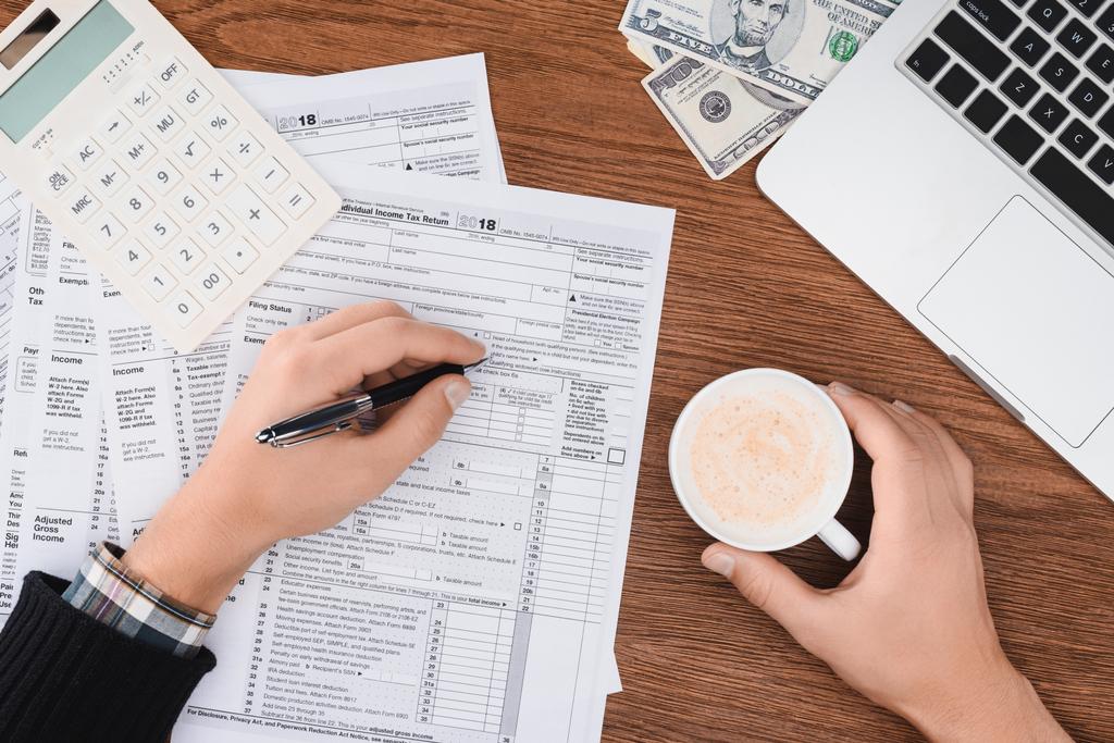 abgeschnittene Ansicht eines Mannes mit Kaffeetasse und dem Ausfüllen von Steuerformularen am Arbeitsplatz - Foto, Bild