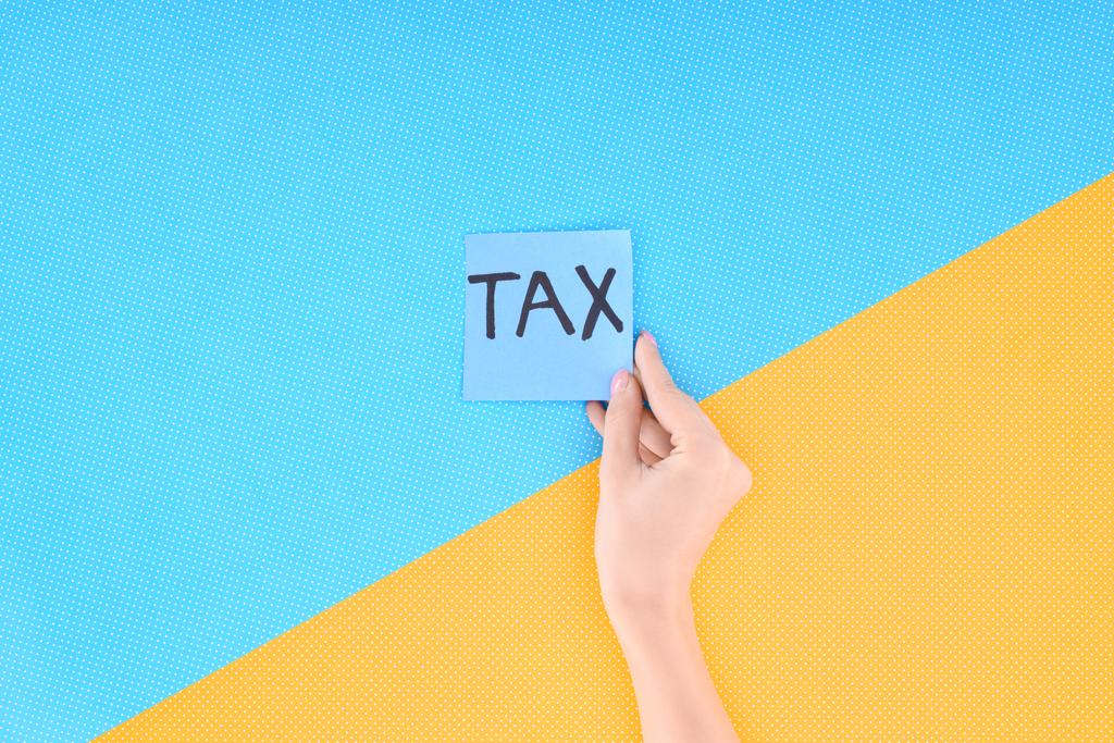 vista recortada de la mujer sosteniendo tarjeta azul con la palabra 'impuesto' sobre fondo azul y amarillo
 - Foto, imagen
