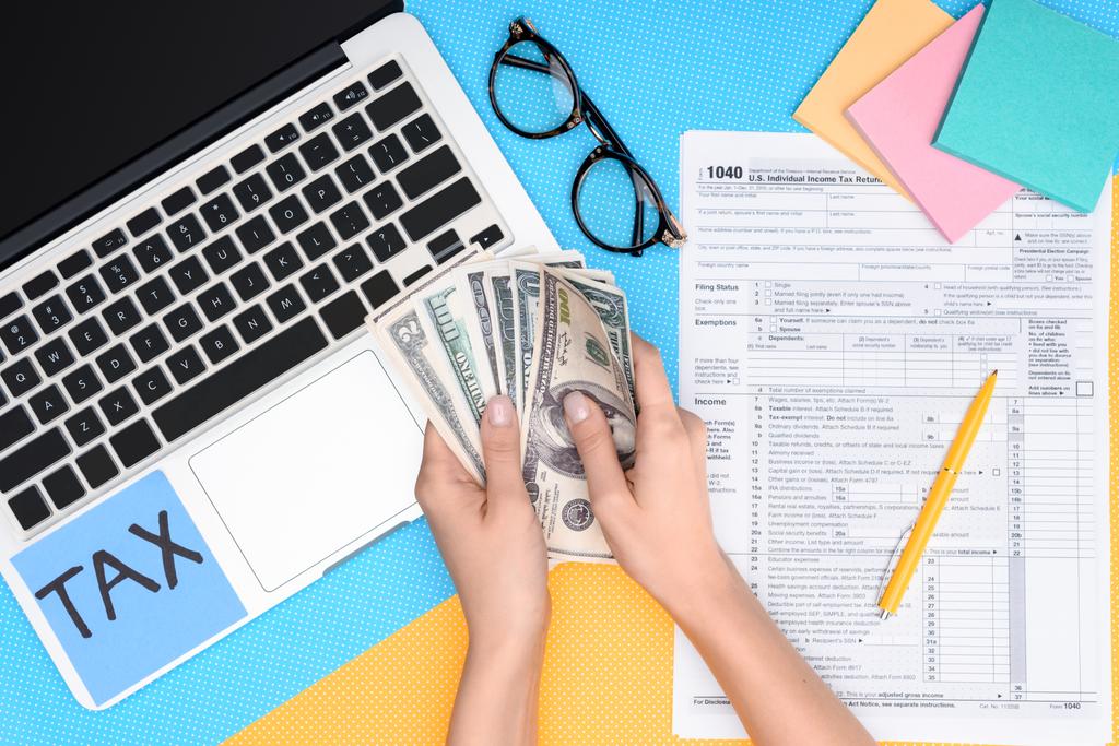 oříznutý pohled ženy držící dolarové bankovky na pracovišti s notebookem a daňové formuláře na pozadí - Fotografie, Obrázek