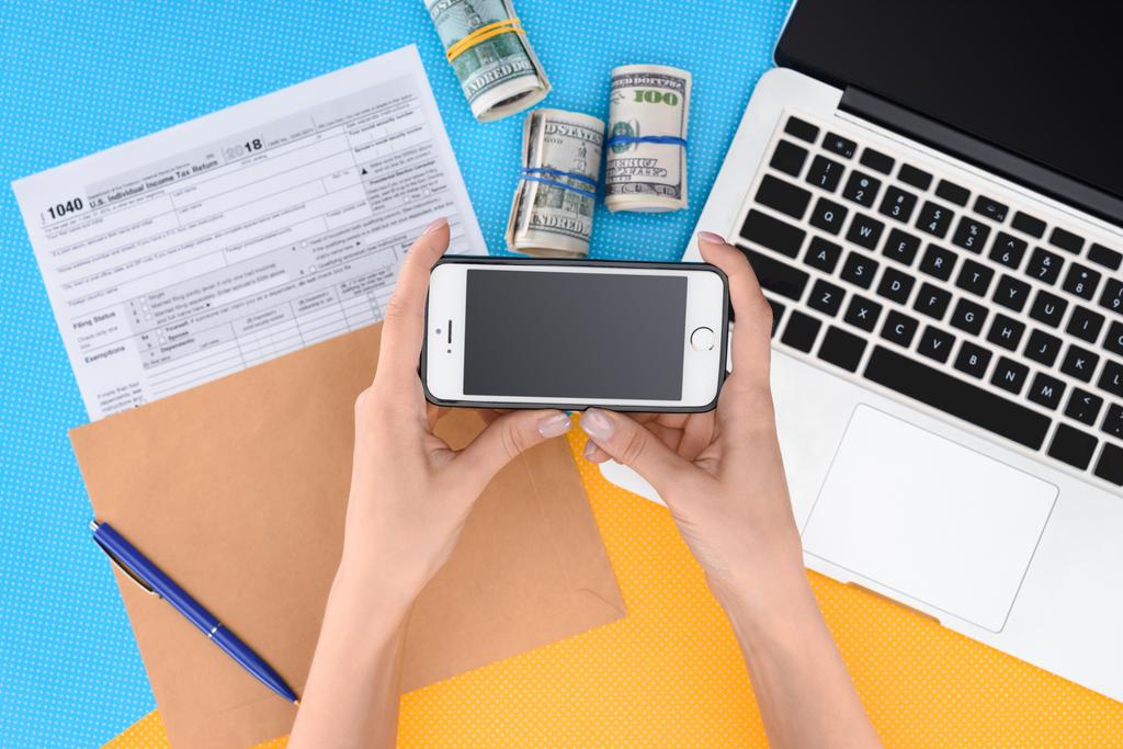 частковий вид жінки, що тримає смартфон з порожнім екраном на робочому місці з ноутбуком, податковою формою та грошовими рулонами на фоні
 - Фото, зображення