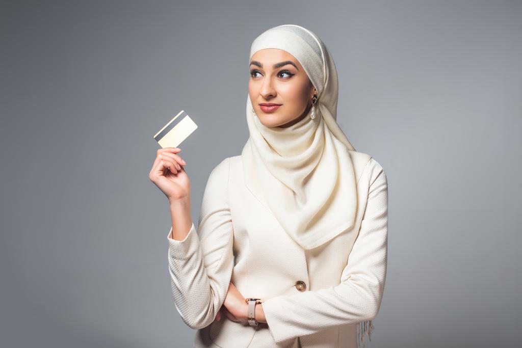 hermosa mujer musulmana sonriente sosteniendo tarjeta de crédito y mirando hacia otro lado aislado en gris
 - Foto, imagen