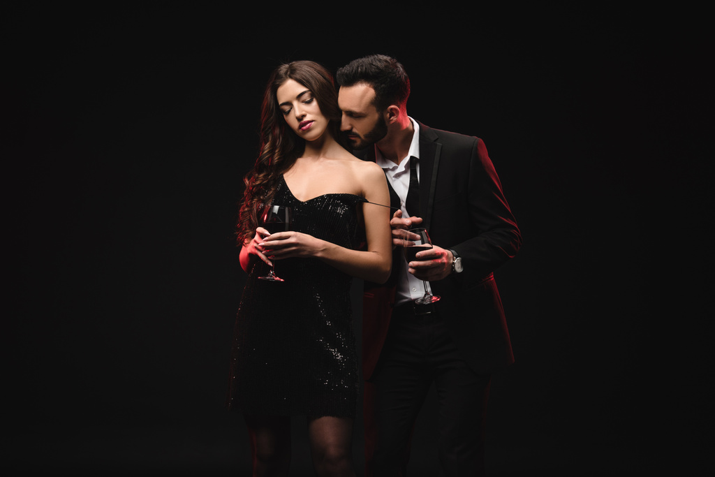 Αισθησιακό ζευγάρι σε μαύρο φόρεμα και σμόκιν κρατώντας γυαλιά με κόκκινο κρασί που απομονώνονται σε μαύρο  - Φωτογραφία, εικόνα