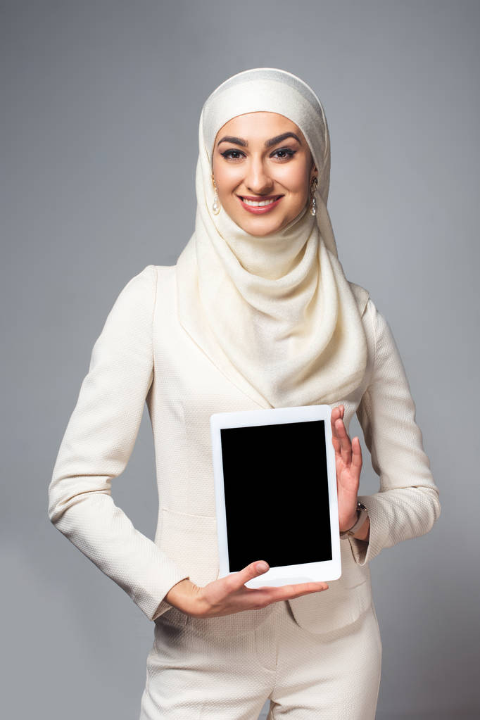fiatal muzulmán nő gazdaság digitális tabletta-val üres képernyő, és mosolyogva kamera elszigetelt szürke - Fotó, kép