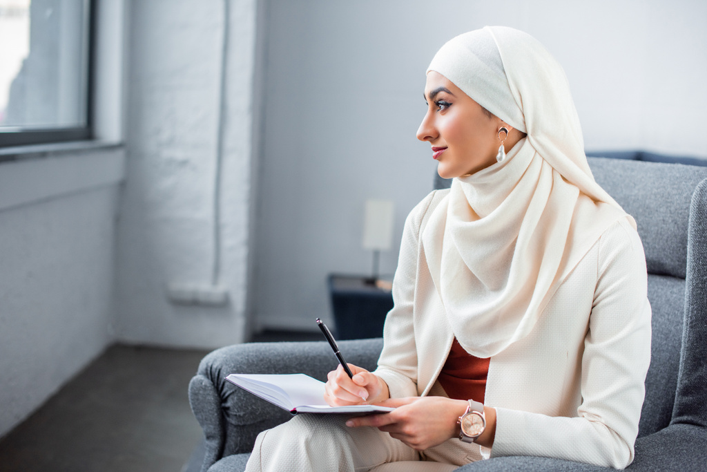 όμορφη νεαρή μουσουλμανική γυναίκα κάθεται στην καρέκλα με το σημειωματάριο και αναζητούν μακριά - Φωτογραφία, εικόνα