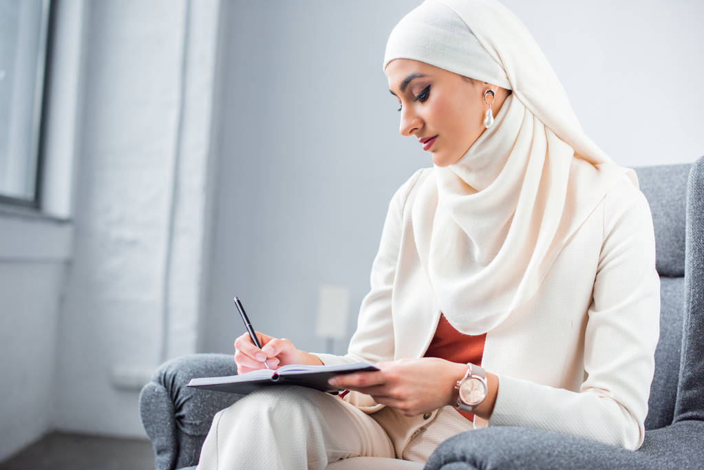 красивая молодая мусульманка сидит в кресле и пишет в блокноте
 - Фото, изображение