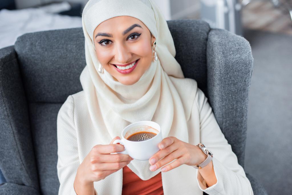 一杯のコーヒーを保持しているとカメラに笑顔の若いイスラム教徒の女性 - 写真・画像