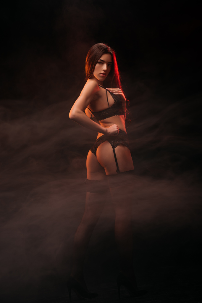 mooie seksuele jonge vrouw in zwarte lingerie poseren in donkere rokerige kamer - Foto, afbeelding