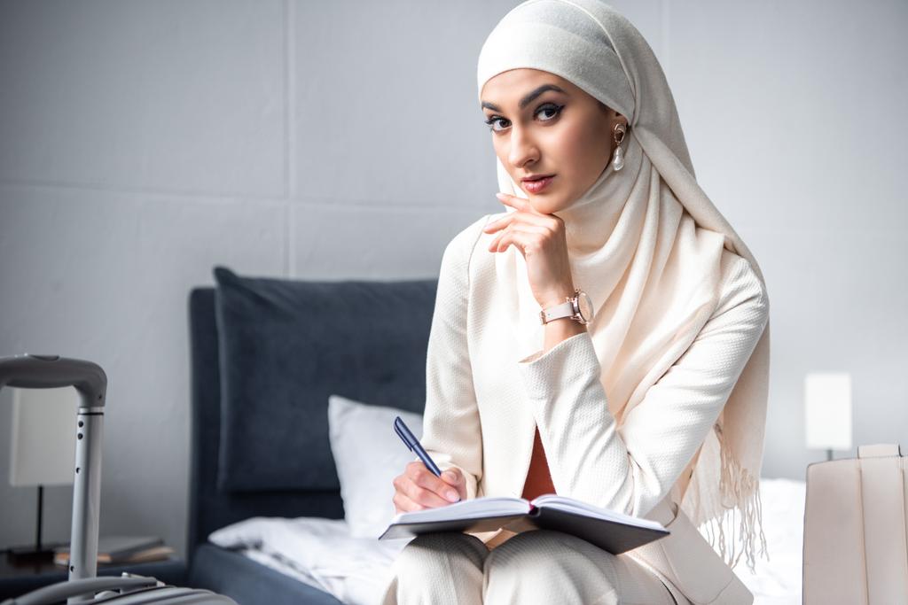 mladá Muslimská žena sedí na posteli a při pohledu na fotoaparát při zápisu v poznámkovém bloku - Fotografie, Obrázek