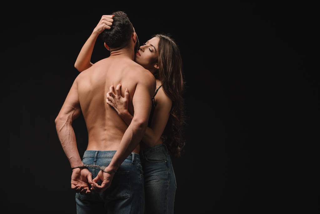 seductive young woman hugging shirtless man in handcuffs isolated on black - Photo, Image