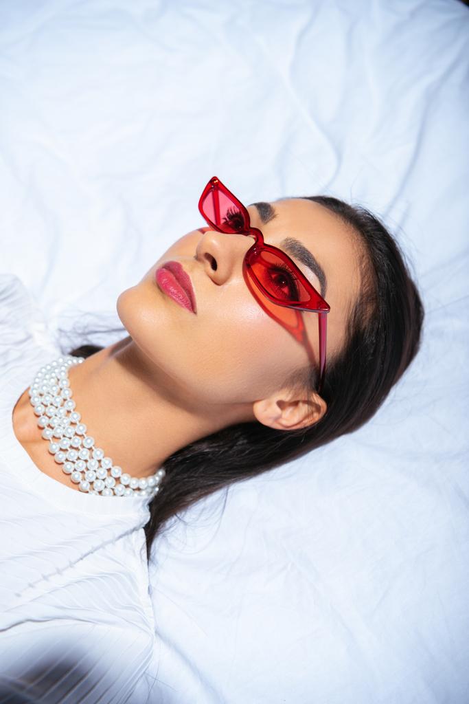 красивая элегантная брюнетка в красных очках лежит на кровати и смотрит вверх
 - Фото, изображение