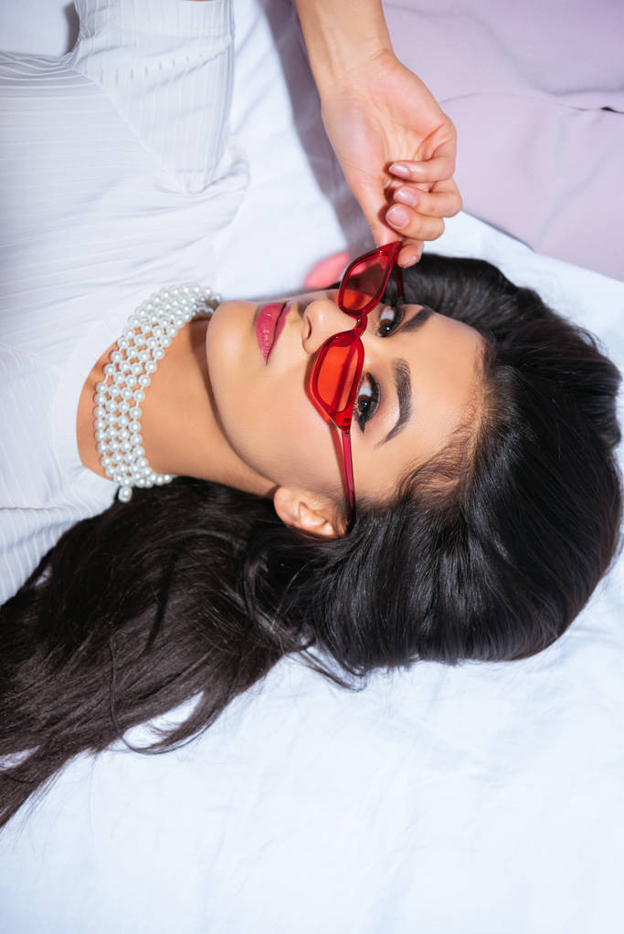 high angle view of elegant brunette girl in red eyeglasses lying on bed and looking at camera - Photo, Image